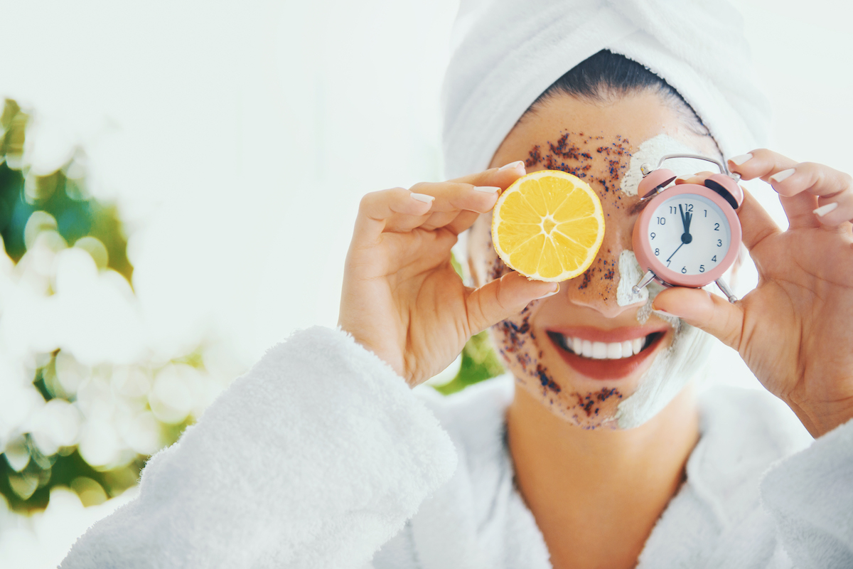 Woman with body scrub on her face and holding an orange slice to cover one eye and a clock timer to cover to other